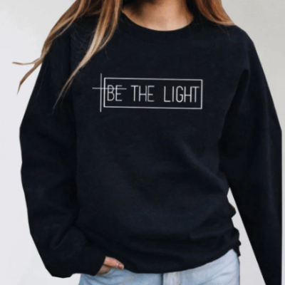 Kvinna bär "Be the Light" sweatshirt. Bekväm, inspirerande damtröja i bomull/polyester för mode och komfort.